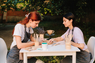 Outdoor pottery session shows students working with clay. Outdoor pottery class. Two women enjoy outdoor pottery class, sitting at table with clay and carving tools while learning ceramic crafting.