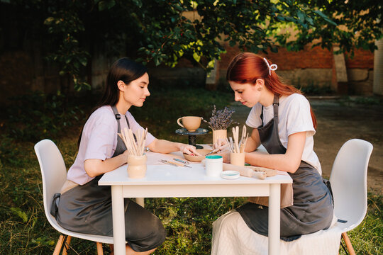 Outdoor pottery class. Two women enjoy outdoor pottery class, sitting at table with clay and carving tools while learning ceramic crafting. Outdoor pottery session shows students working with clay.