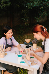 Outdoor pottery class. Two women enjoy outdoor pottery class, sitting at table with clay and carving tools while learning ceramic crafting. Outdoor pottery session shows students working with clay.