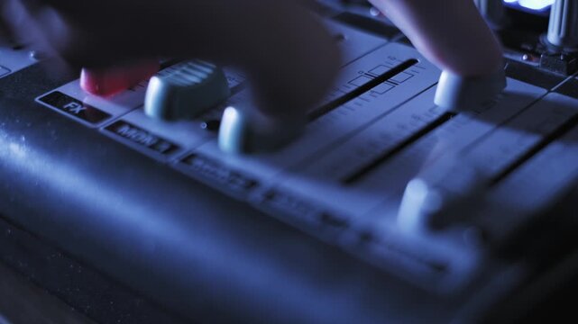 Macro shot of a finger pushing the main volume fader on an audio mixing console, showing channel labels like MAIN 1 and MON 2 under dynamic, shifting purple and orange club lights in a dark studio.