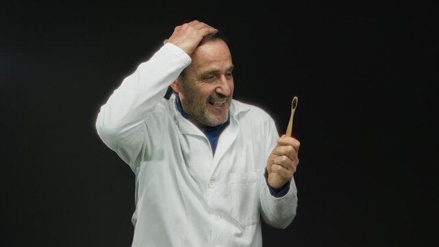 Man in doctor coat holding bamboo toothbrush against black background expresses surprise with hand gestures.