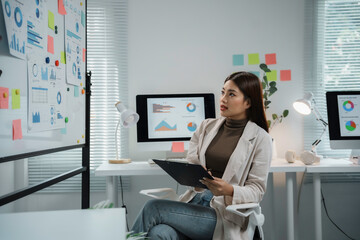 Young Asian businesswoman working in office, holding a clipboard and looking at charts and graphs...