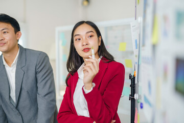 Young Asian businesswoman in red blazer presenting ideas while holding a marker, collaborating with colleagues during a busy office meeting