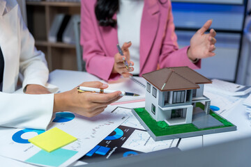 Businesswomen meeting about property investment, showing a house model and charts on a desk during a negotiation