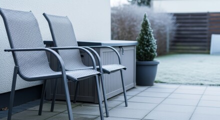 Gray wicker chairs and storage box on residential terrace covered with frost during first snowfall. Cold winter outdoor living space with evergreen plant.