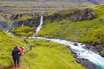 Fototapeta premium Group of people hiking towards a waterfall with a running stream and mountains