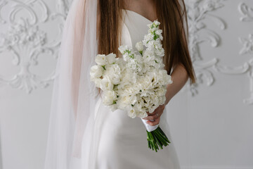 Bride in a white satin wedding dress holding a lush, vertical white bouquet of stock flowers and eustoma carnations against a background of classic white stucco wall paneling