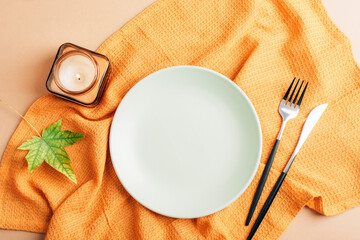 Autumn table arrangement with plate, burning candle, cutlery and yellow napkin on beige table. Top view, flat lay, mockup