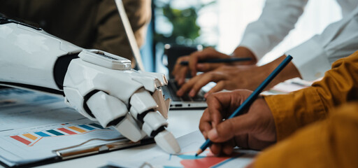 A close-up view of a futuristic robotic hand working alongside humans, illustrating innovative collaboration in a modern workspace with various tools and data analysis charts.Noogenesis