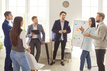 Obraz premium Young business man talking about work project for a group of company employees standing in meeting room. Confident business people discussing company growth on a conference in modern office.