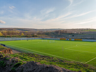 A bright green soccer field sits on a gentle slope, surrounded by hills and trees. The sunlight creates a warm atmosphere, making it an inviting space for local players. © Ion B