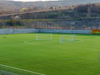 Naklejka premium A soccer field sits empty under a clear sky with two goals at either end. The bright green grass contrasts with the surrounding hills, showcasing a peaceful afternoon scene.