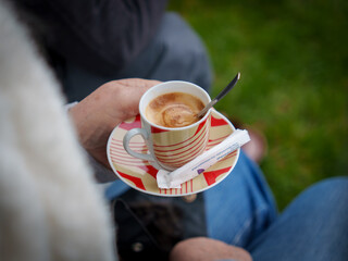 A person holds a cup of coffee with a patterned saucer while sitting in a park. The scene captures a sense of relaxation and enjoyment during a pleasant day outside.