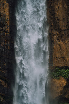 View of a powerful waterfall cascading down rugged cliffs, with water's mist and greenery creating a serene scene, Kapas Biru Waterfall, Jawa Timur, Indonesia.