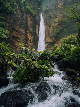 View of a cascading waterfall plunges amidst verdant foliage and rugged cliffs, creating a serene scene of nature's grandeur, Kapas Biru Waterfall, Jawa Timur, Indonesia.