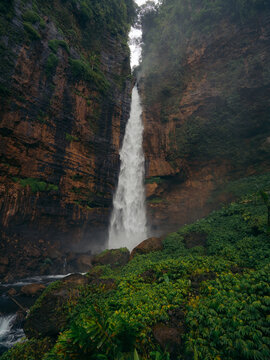 View of a powerful waterfall cascading down between towering rock faces, embraced by lush green vegetation, creating a scene of raw natural beauty, Kapas Biru Waterfall, Jawa Timur, Indonesia.