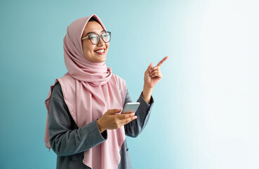 Smiling woman in hijab holds smartphone and points finger to side. She wears glasses and a grey jacket. Modern communication and tech lifestyle concept.