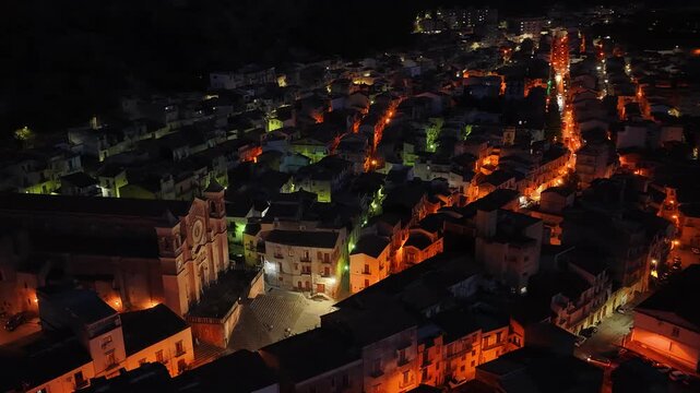 Aerial drone night view of Collesano, a historic Sicilian mountain town at dusk with Basilica San Pietro church and illuminated stone streets in the Madonie Mountains, Sicily, Italy