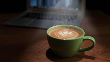 Green mug of latte with latte art next to a laptop on a wooden table
