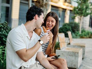Happy couple  eating a pastry croissant, smiling and looking away on a city street