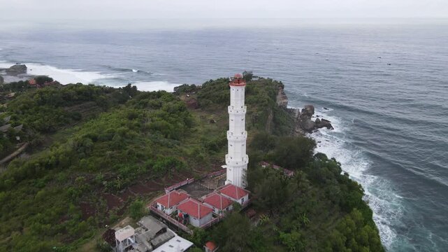 Aerial view of a stark white lighthouse piercing through the lush greenery against the turbulent ocean waves, Baron, Yogyakarta, Indonesia.