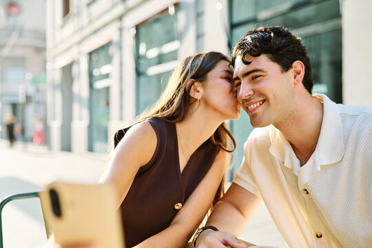 Young couple enjoying conversation and coffee together and taking a selfie with a smartphone at an outdoor urban cafe table