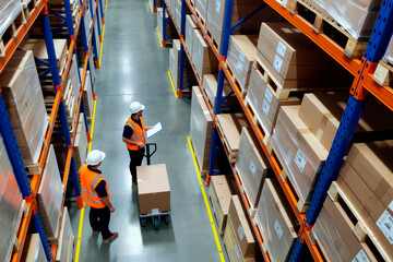 Two middle aged Caucasian men wearing safety vests and helmets working in warehouse, one holding clipboard and checking inventory while other moving box on hand truck between shelves