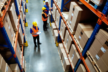 Three middle aged workers, two men and one woman, wearing safety helmets and vests, inspecting inventory and taking notes while standing between tall shelves in warehouse storage area