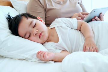 Asian child sleeping peacefully on bed with head resting on pillow while adult woman using digital tablet in background, close up showing relaxed facial expression and gentle posture