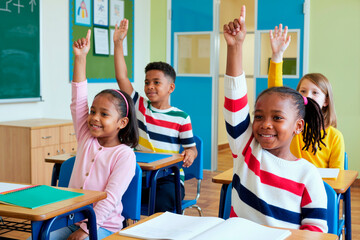 Group of multiethnic children sitting at desks raising hands in classroom, smiling and engaging with teacher, school supplies and notebooks on desks, learning environment visible