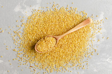 Golden bulgur wheat grains scattered on a textured gray surface, with a wooden spoon filled with bulgur, showcasing natural ingredients for healthy cooking and meal preparation
