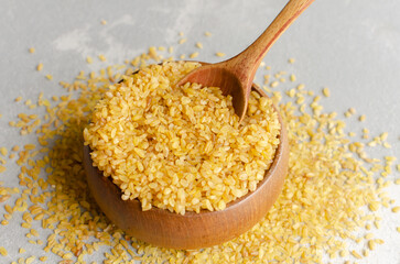 Yellow bulgur wheat in a wooden bowl with a wooden spoon, surrounded by scattered grains on a light textured surface, showcasing healthy grains for culinary use