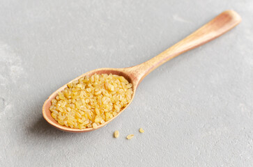 Wooden spoon filled with yellow bulgur grain rests on a textured gray surface, showcasing the natural color and texture of this nutritious ingredient for culinary use