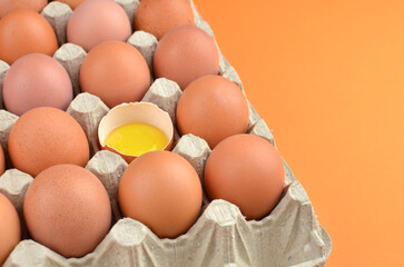 Fresh brown eggs in a carton with one cracked shell revealing bright yellow yolk, set against a vibrant orange background, showcasing natural food elements and textures
