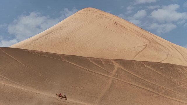 Aerial view of camels traversing the vast, undulating sand dunes, their trails etching patterns into the landscape's golden hues, Turpan, Xinjiang, China.