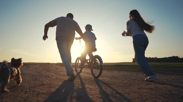 Family on dirt road at sunset. Family teaches their son how to ride bike. A father and child are riding a bicycle. A family on a gravel road during lifestyle sunset.