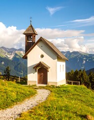 Small, light-cream-colored chapel nestled in a grassy mountainside, with a gravel path leading to it.  Wooden bell tower.  Blue sky with puffy clouds, and distant mountains.  Alpine scenery