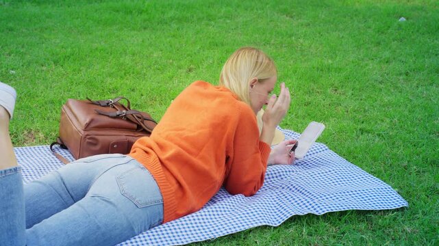 Young blonde woman relaxing on checkered blanket in park, lying on stomach on green grass while engrossed in reading an e-book on her digital tablet during a sunny day