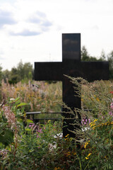 A Black Granite Cross Gravestone On An Overgrown Grave With Wildflowers In A Cemetery. Vertical Shot, Concept Of Death And Memory.