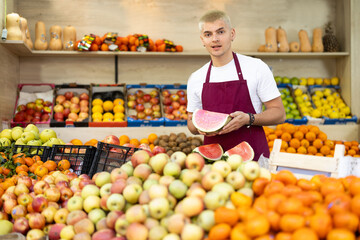 Young guy seller in apron offers fresh watermelon at vegetable market