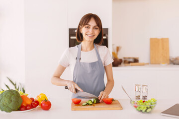 Happy young lady cooking fresh vegetable salad, chopping cucumber and smiling at camera in kitchen. Woman preparing lunch at home, enjoying eating healthy food, copy space
