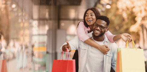 Portrait of smiling African American man giving piggyback ride to his happy woman who holding shopper bags, couple looking and pointing at shop window, walking near shopping center