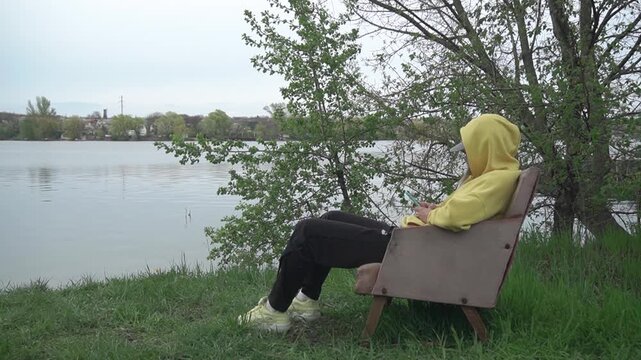 a girl bright yellow hoodie hood and cap looks intently phone while sitting in an old vintage chair bank river with a fast current, green grass around and the wind sways the trees, a cool day