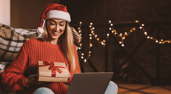 Winter Holidays, Virtual Celebration. Smiling woman in santa claus hat opening wrapped gift box at home in front of laptop video-calling with distant relatives friends, sitting on floor in living room