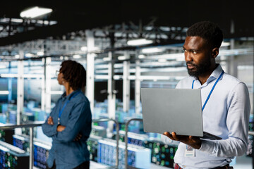 Fototapeta premium Smiling black technician and colleague in data center reviewing large language model visualizations on PC. Professional IT engineering team analyzing neural network performance outputs.