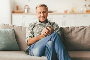 Fototapeta premium Senior Male Using Smartphone With New Application Texting, Networking In Social Media Sitting On Sofa At Home. Great Application, People And Gadgets, Retirement Lifestyle Concept