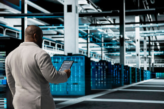 Technician walking in data center running lines of code on tablet, troubleshooting rackmounts. African american IT specialist using programming scripts on device in server room - Powered by Adobe