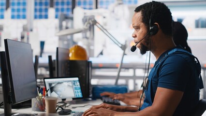 Call center technician handling maintenance inquiries in solar factory, working in customer service industrial tech support. African american operator on headset with clients. Camera A. © DC Studio