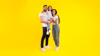 Happy Middle Eastern Family Posing Together, Young Parents Hugging Their Daughter Over Yellow Studio Background. Successful Parenting Concept. Vertical, Full Length Shot
