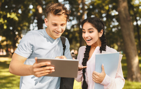 University lifestyle concept. Student guy showing digital tablet to girlfriend, reading news or looking at test results while walking outdoors in college campus